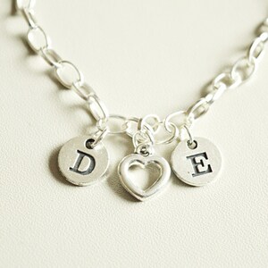 Puede incluir: Un collar de cadena de plata con tres colgantes: un corazón, un colgante con la letra D y un colgante con la letra E.