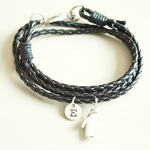 Op de afbeelding: Een zwarte leren gevlochten armband met een zilveren bedel met de letter 'E' erop en een zilveren pingu&iuml;nbedel.