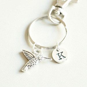 May include: Silver hummingbird keychain with a silver letter K charm. The keychain is attached to a silver key ring.