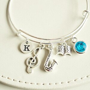 Pode incluir: Pulseira de prata com um charme de nota musical, um saxofón, um livro, uma pedra preciosa azul e um charme com a letra K.