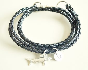 Könnte beinhalten: Ein geflochtenes Armband aus schwarzem Leder mit einem silbernen Hai-Anhänger und einem silbernen Initialen-Anhänger.