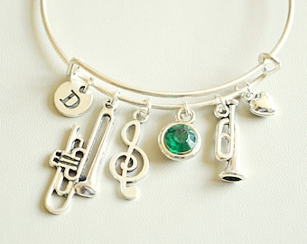 May include: Silver charm bracelet with a green gem, a treble clef, a heart, and two trumpet charms. The bracelet also has a charm with the letter "D" on it.