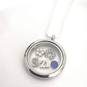 Puede incluir: Collar con un colgante de medall&oacute;n plateado adornado con una gema azul, un dije de herradura, un dije de flor y las letras "S" y "21" con diamantes de imitaci&oacute;n.