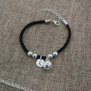 May include: Black braided ankle bracelet with silver beads and charms. One charm is a silver soccer ball and the other is a silver disc with the letter K.