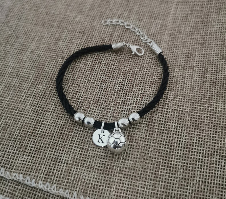 May include: Black braided ankle bracelet with silver beads and charms. One charm is a silver soccer ball and the other is a silver disc with the letter K.