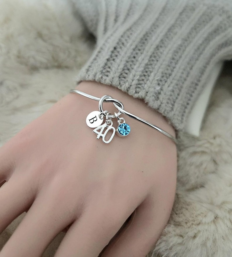 May include: Silver bangle bracelet with charms. The charms include the letter "B", the number "40", and a blue crystal. The bracelet is shown on a person's wrist.