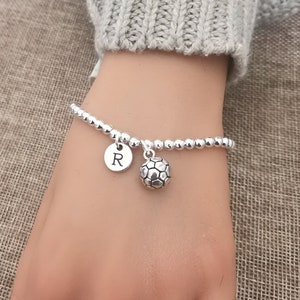 May include: A silver beaded bracelet with a soccer ball charm and a round charm with the letter "R". The bracelet is on a wrist, with a gray sweater in the background.