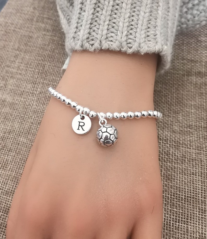 May include: A silver beaded bracelet with a soccer ball charm and a round charm with the letter "R". The bracelet is on a wrist, with a gray sweater in the background.