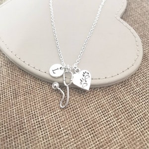 Peut inclure: Collier argent&eacute; avec trois breloques : un cercle avec la lettre "L", un st&eacute;thoscope et un c&oelig;ur avec des empreintes de pattes. Le collier est pr&eacute;sent&eacute; sur une surface grise claire.