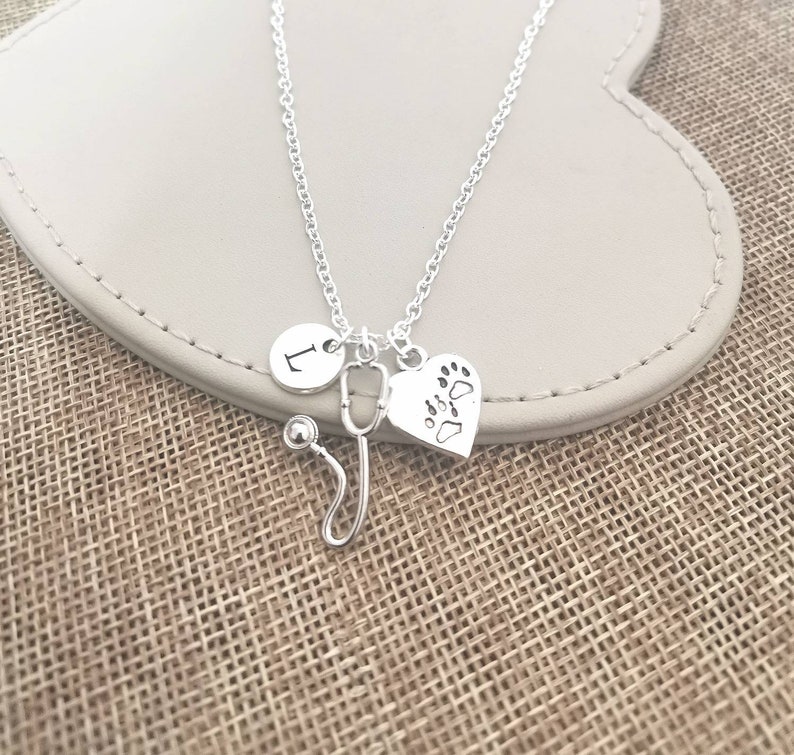 Peut inclure: Collier argent&eacute; avec trois breloques : un cercle avec la lettre "L", un st&eacute;thoscope et un c&oelig;ur avec des empreintes de pattes. Le collier est pr&eacute;sent&eacute; sur une surface grise claire.