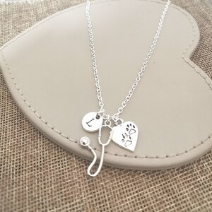Peut inclure: Collier argent&eacute; avec un charme en forme de st&eacute;thoscope, un charme en forme de c&oelig;ur avec des empreintes de pattes et un charme rond avec la lettre "L". Le collier est pr&eacute;sent&eacute; sur une surface en forme de c&oelig;ur, gris clair.