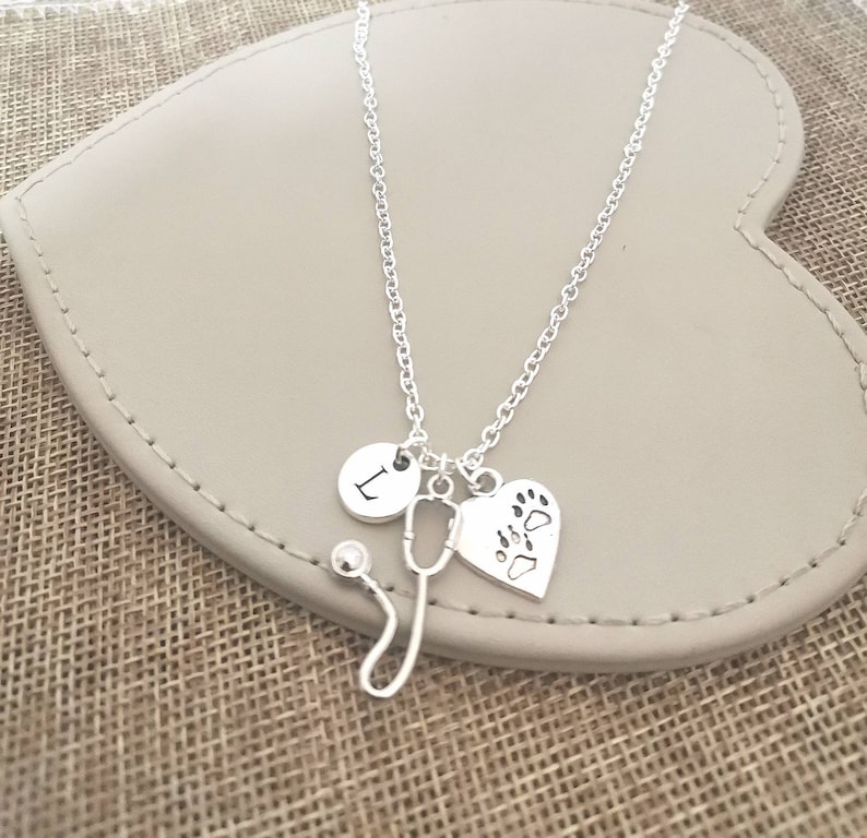 Peut inclure: Collier argent&eacute; avec un charme en forme de st&eacute;thoscope, un charme en forme de c&oelig;ur avec des empreintes de pattes et un charme rond avec la lettre "L". Le collier est pr&eacute;sent&eacute; sur une surface en forme de c&oelig;ur, gris clair.