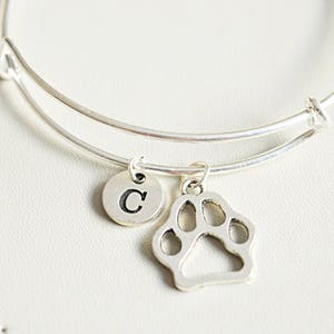 Puede incluir: Pulsera de plata con un dije de huella de pata y un dije con la letra C.