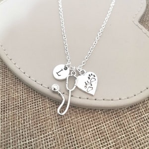 Peut inclure: Collier argent&eacute; avec un charme en forme de st&eacute;thoscope, un charme en forme de c&oelig;ur avec une empreinte de patte et un charme rond avec la lettre "I". Le collier est pr&eacute;sent&eacute; sur une surface gris clair.