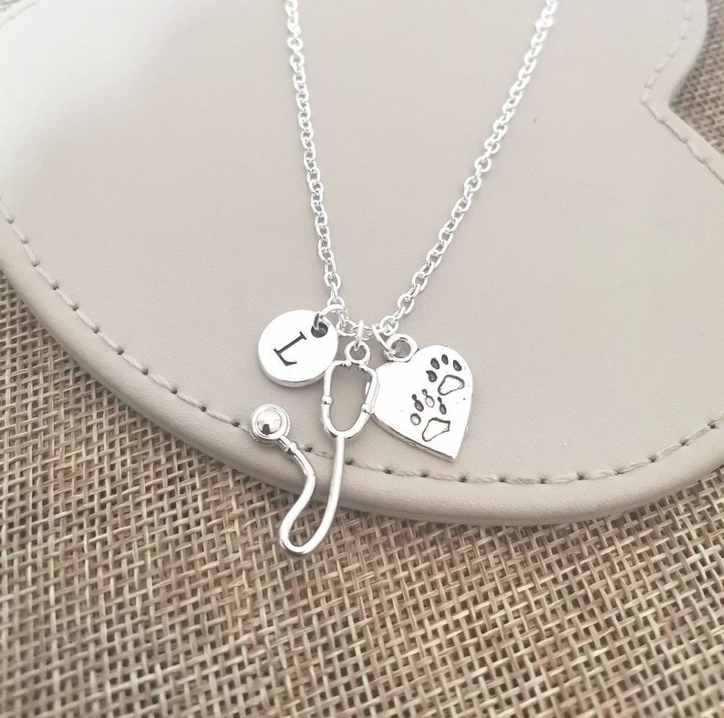 Peut inclure: Collier argent&eacute; avec un charme en forme de st&eacute;thoscope, un charme en forme de c&oelig;ur avec une empreinte de patte et un charme rond avec la lettre "I". Le collier est pr&eacute;sent&eacute; sur une surface gris clair.