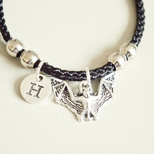 May include: Black braided cord bracelet with silver beads and a silver bat charm. The bat charm has a small silver disc with the letter "H" on it.