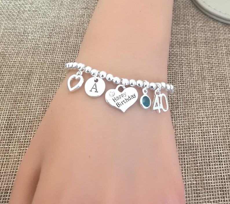 May include: Silver charm bracelet with a heart charm that says "Happy Birthday", a charm with the letter "A", a charm with a turquoise stone, and a charm with the number "40".