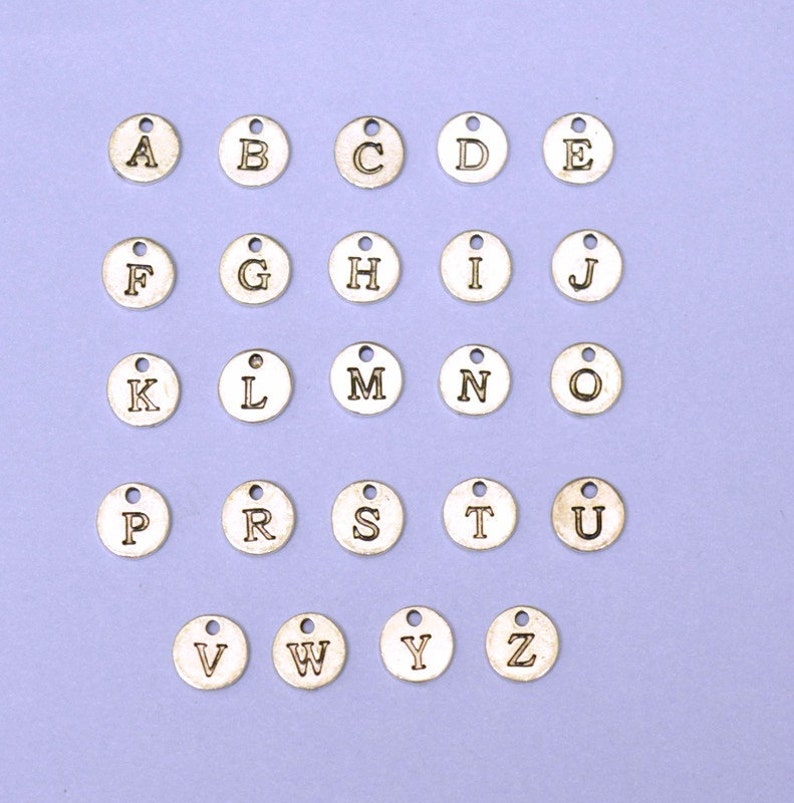 May include: A set of 26 silver-toned metal alphabet letter charms. Each charm is round and features a single letter of the alphabet in a black font.