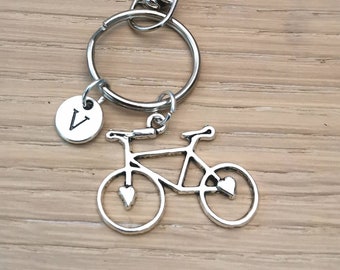 May include: Silver-toned bicycle charm keychain with a round tag featuring the letter "V". The bicycle has heart-shaped details on the wheels. The keychain includes a clasp and a split ring for attaching keys.