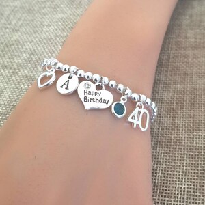 May include: Silver charm bracelet with a heart charm, a letter A charm, a charm that says "Happy Birthday", a green gemstone charm, and a charm with the number 40.