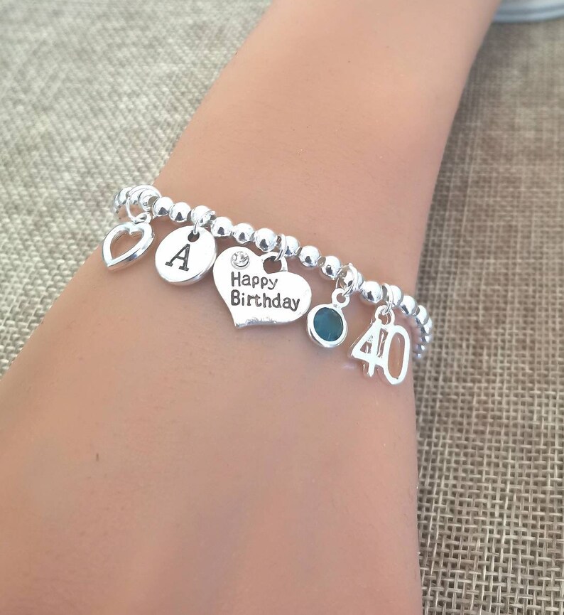 May include: Silver charm bracelet with a heart charm, a letter A charm, a charm that says "Happy Birthday", a green gemstone charm, and a charm with the number 40.