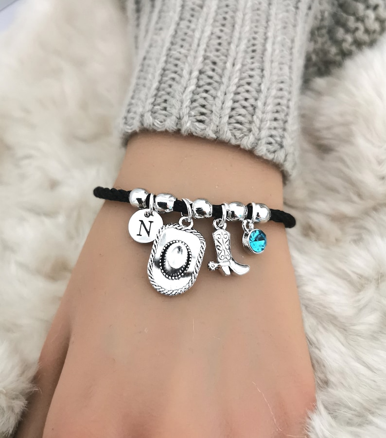 May include: A black elastic bracelet with silver charms, including a cowboy hat, a cowboy boot, a letter 'N' charm, and a blue crystal charm.