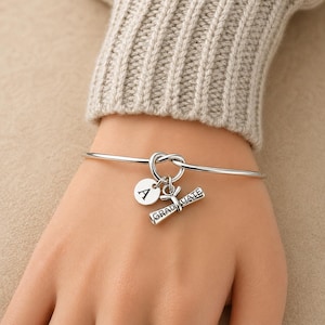 May include: A silver-toned bangle bracelet with a knot design. It features a round charm with the letter "A" and a graduation scroll charm that reads "GRADUATE". The bracelet is worn on a wrist.
