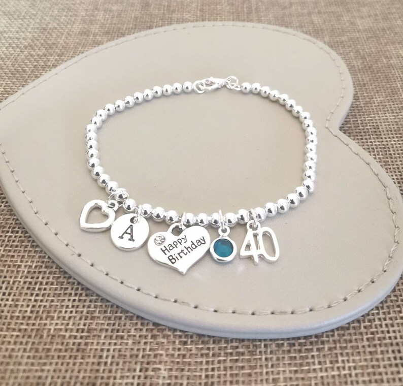 May include: Silver charm bracelet with a heart charm, a charm with the letter A, a heart charm with the words "Happy Birthday", a blue gemstone charm, and a charm with the number 40.