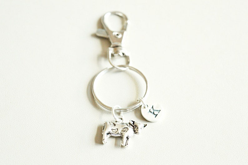 May include: Silver cow keychain with a silver ring and a silver clip. The keychain also has a silver charm with the letter "R" on it.