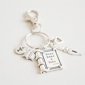 Op de afbeelding: Zilverkleurige sleutelhanger met diverse bedels. Bedels zijn onder meer een boek, een rol met de tekst "Once Upon a Time", een pen, een hart en de letter "T". De sleutelhanger heeft een sluiting.