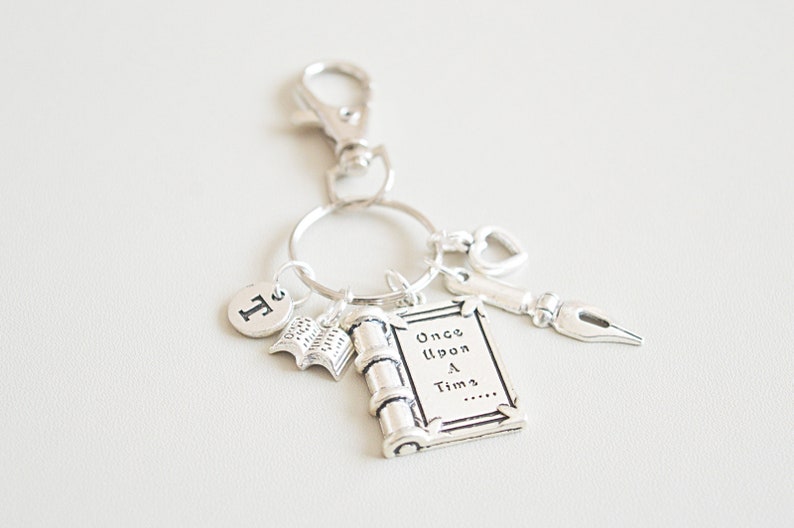 Op de afbeelding: Zilverkleurige sleutelhanger met diverse bedels. Bedels zijn onder meer een boek, een rol met de tekst "Once Upon a Time", een pen, een hart en de letter "T". De sleutelhanger heeft een sluiting.