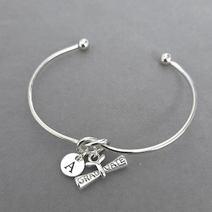 May include: Silver-toned bangle bracelet with a graduation theme. The bracelet features a charm with the letter "A", a graduation scroll charm, and the word "GRADUATE". The bracelet is a simple, elegant design.