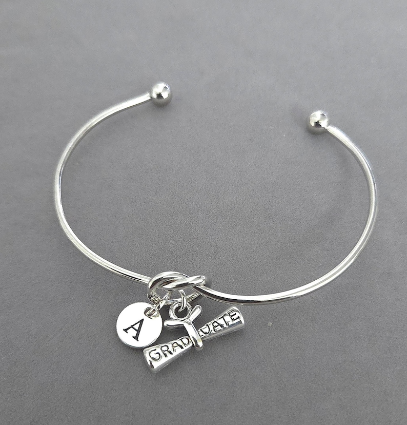 May include: Silver-toned bangle bracelet with a graduation theme. The bracelet features a charm with the letter "A", a graduation scroll charm, and the word "GRADUATE". The bracelet is a simple, elegant design.