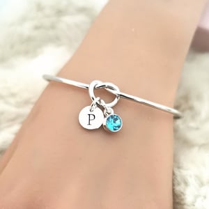 May include: A silver knot bangle bracelet with a small silver charm with the letter "P" and a light blue crystal charm.