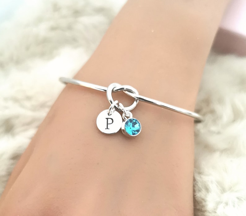 May include: A silver knot bangle bracelet with a small silver charm with the letter "P" and a light blue crystal charm.