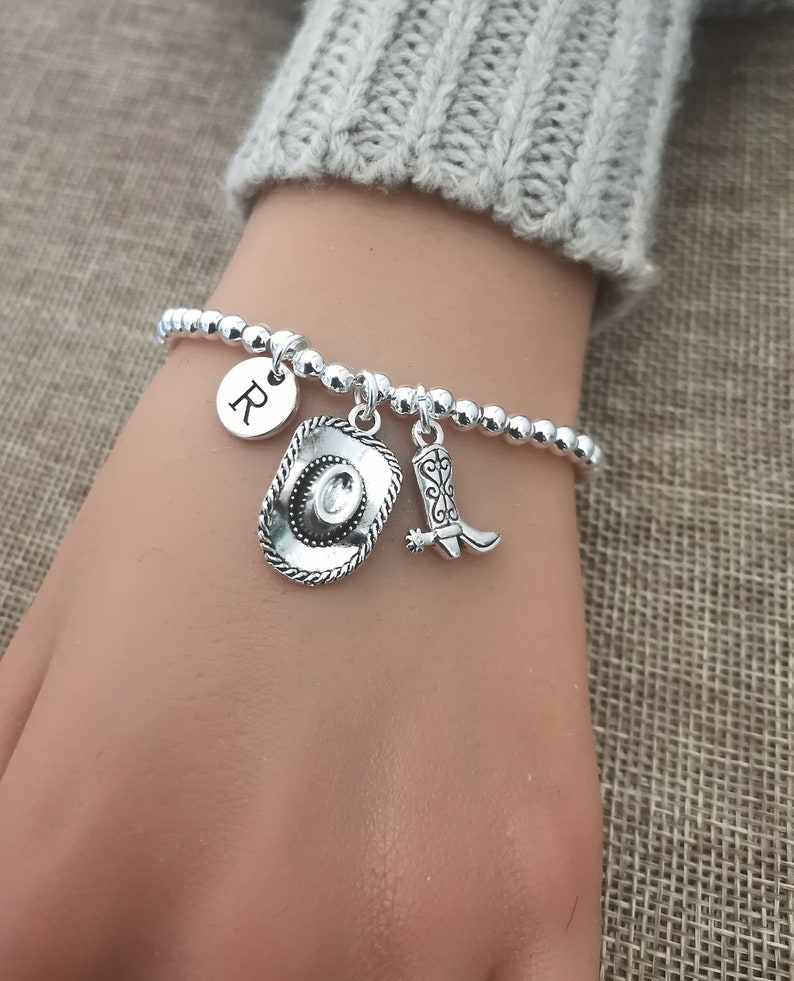 May include: Silver beaded bracelet with a cowboy hat charm, a cowboy boot charm, and a round charm with the letter "R". The bracelet is on a wrist, with a gray sweater in the background.