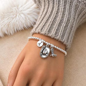 May include: A silver beaded bracelet with three charms: a round charm with the letter "A", a cowboy hat, and a cowboy boot. The bracelet is worn on a wrist, with a neutral-colored sweater in the background.