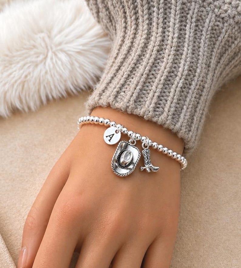 May include: A silver beaded bracelet with three charms: a round charm with the letter "A", a cowboy hat, and a cowboy boot. The bracelet is worn on a wrist, with a neutral-colored sweater in the background.