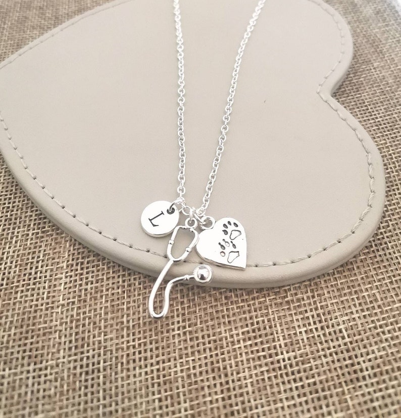 Peut inclure: Un collier argent&eacute; avec trois breloques : un st&eacute;thoscope, un c&oelig;ur avec des empreintes de pattes et un cercle avec la lettre "L". Le collier est pr&eacute;sent&eacute; sur une surface en forme de c&oelig;ur, de couleur gris clair.