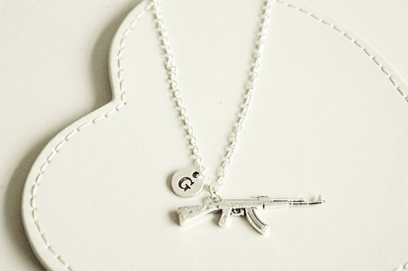 May include: A silver necklace with a charm of a small silver gun and a silver disc with the letter "G" on it.