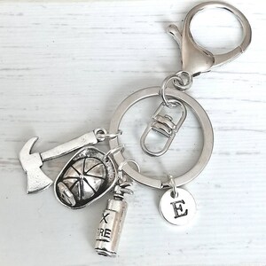 May include: Silver keychain with a firefighter theme. It features a small axe, a firefighter's helmet, a fire extinguisher, and a charm with the letter "E".