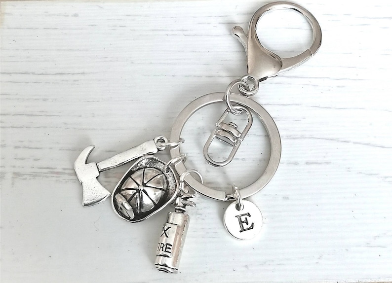 May include: Silver keychain with a firefighter theme. It features a small axe, a firefighter's helmet, a fire extinguisher, and a charm with the letter "E".