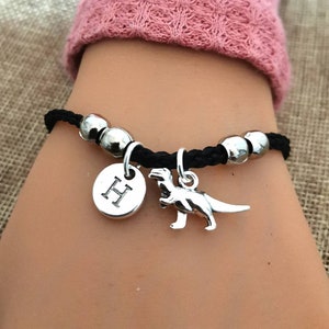 May include: Black braided bracelet with silver beads and charms. The charms include a silver dinosaur and a silver disc with the letter "H" on it.