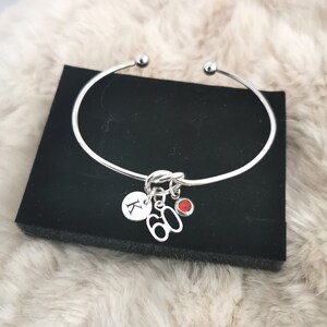 K&ouml;nnte beinhalten: Silbernes Armreif-Armband mit einem Anh&auml;nger, auf dem "60" steht, und einem kleinen roten Strassstein. Das Armband hat auch einen Anh&auml;nger mit dem Buchstaben "K" darauf.
