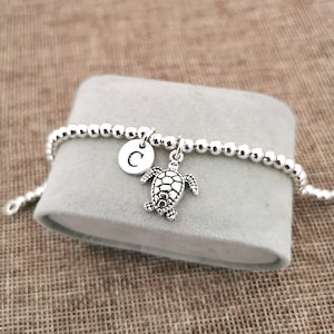 Könnte beinhalten: Silbernes Perlenarmband mit einem silbernen Schildkrötenanhänger und einem silbernen Scheibenanhänger mit dem eingravierten Buchstaben "C".