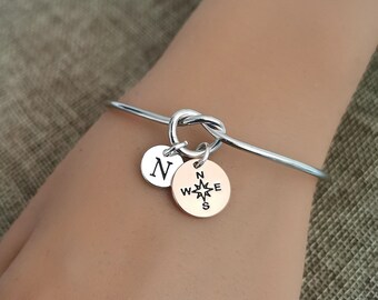 Puede incluir: Un brazalete de nudo de plata con un dije de brújula y un dije con la letra N. El brazalete está en la muñeca de una persona.