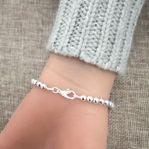 May include: A silver beaded bracelet with a lobster clasp is shown on a wrist. The bracelet is made of small, round, shiny beads. The wrist is partially visible, and the person is wearing a gray sweater.