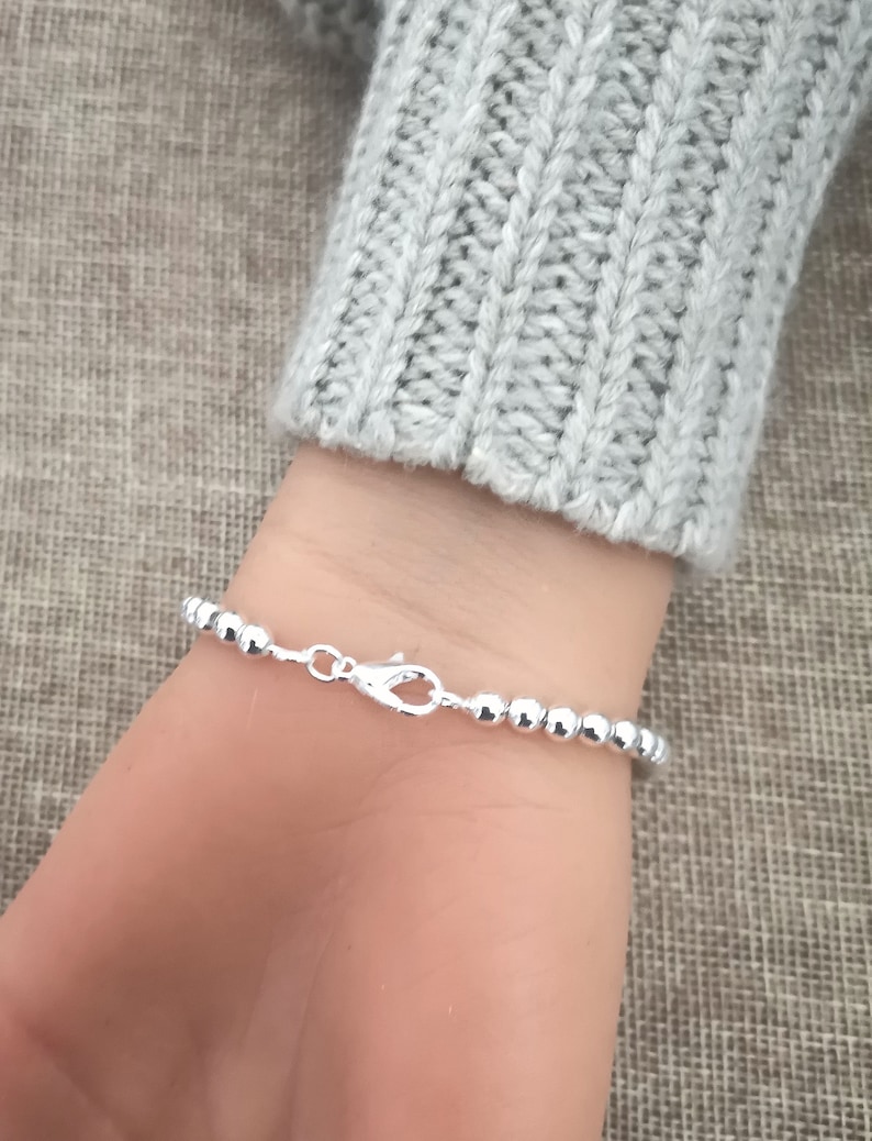 May include: A silver beaded bracelet with a lobster clasp is shown on a wrist. The bracelet is made of small, round, shiny beads. The wrist is partially visible, and the person is wearing a gray sweater.