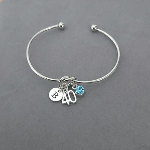 May include: Silver bangle bracelet with a knot design. It has three charms: a round charm with the letter "B", the number "40", and a blue crystal. The bracelet is presented on a grey surface.