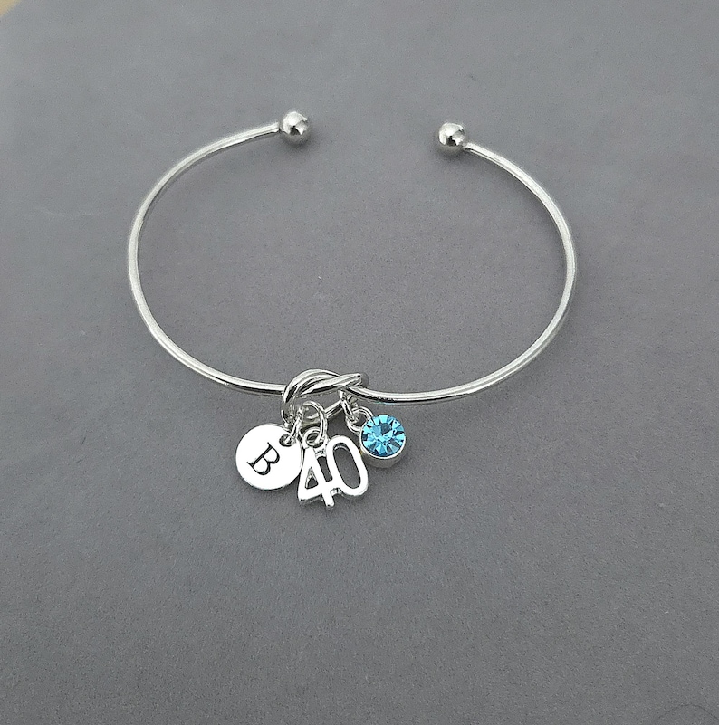 May include: Silver bangle bracelet with a knot design. It has three charms: a round charm with the letter "B", the number "40", and a blue crystal. The bracelet is presented on a grey surface.